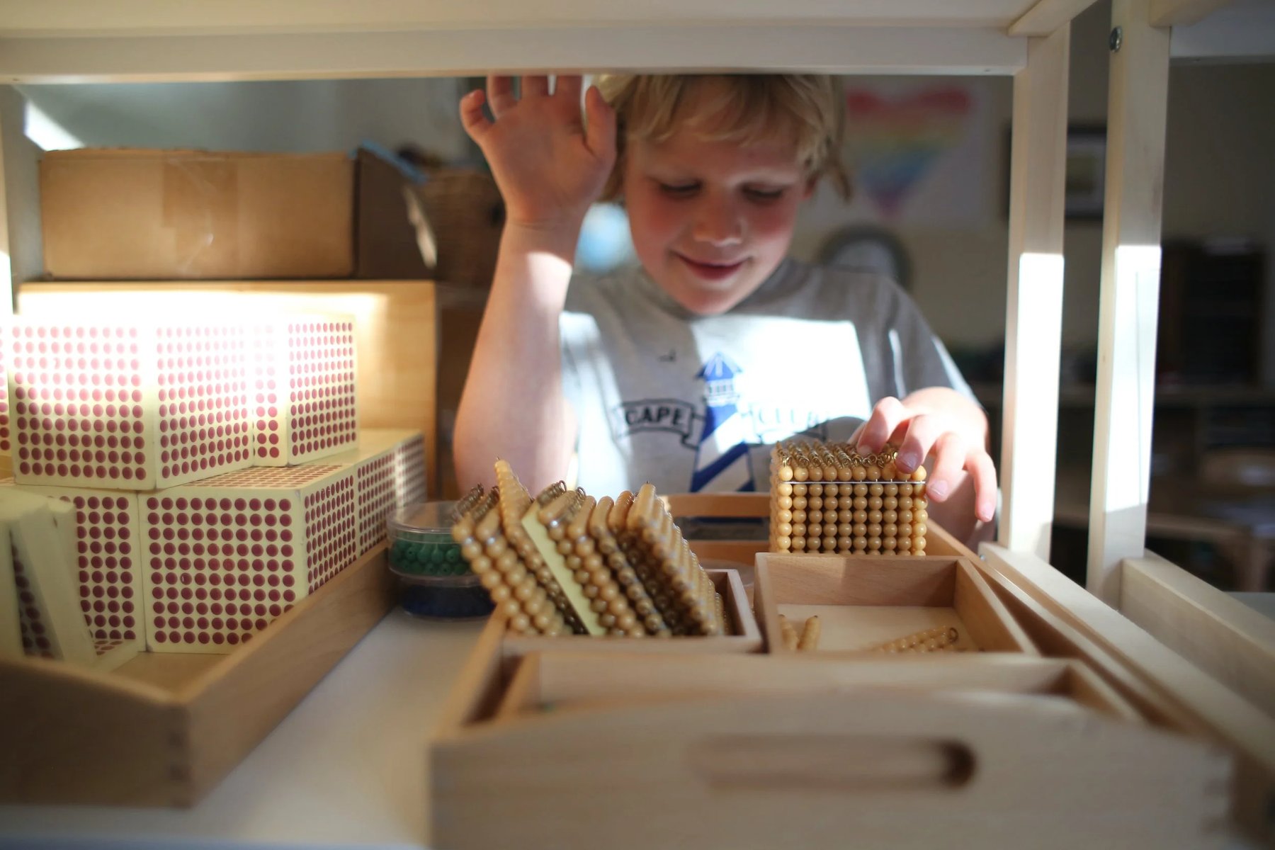 A smiling blond boy reaches through wooden classroom shelving toward trays of golden Montessori bead materials