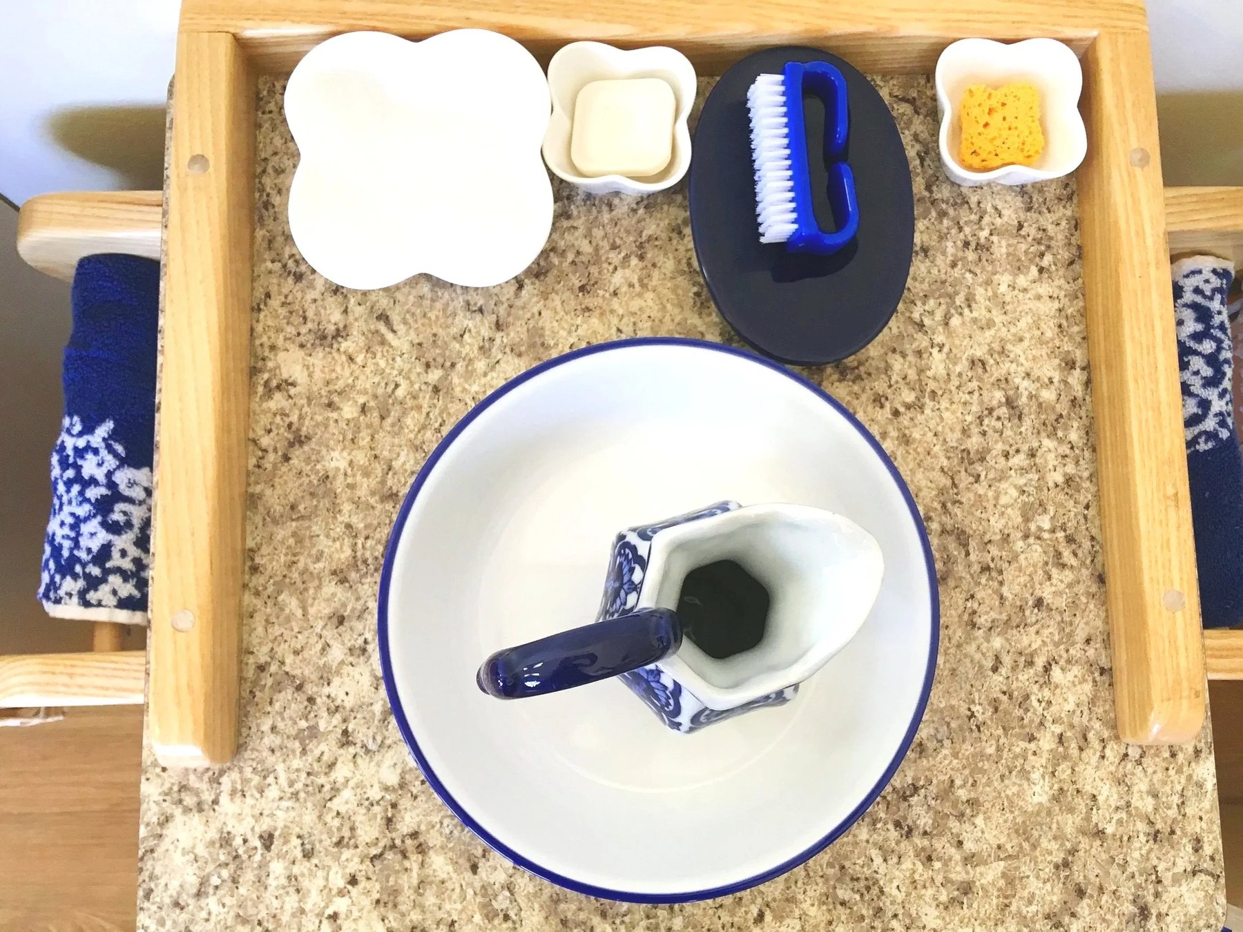 Montessori practical-life handwashing setup on a wooden stand with a basin, decorated pitcher, soap and brush dishes, and patterned towels
