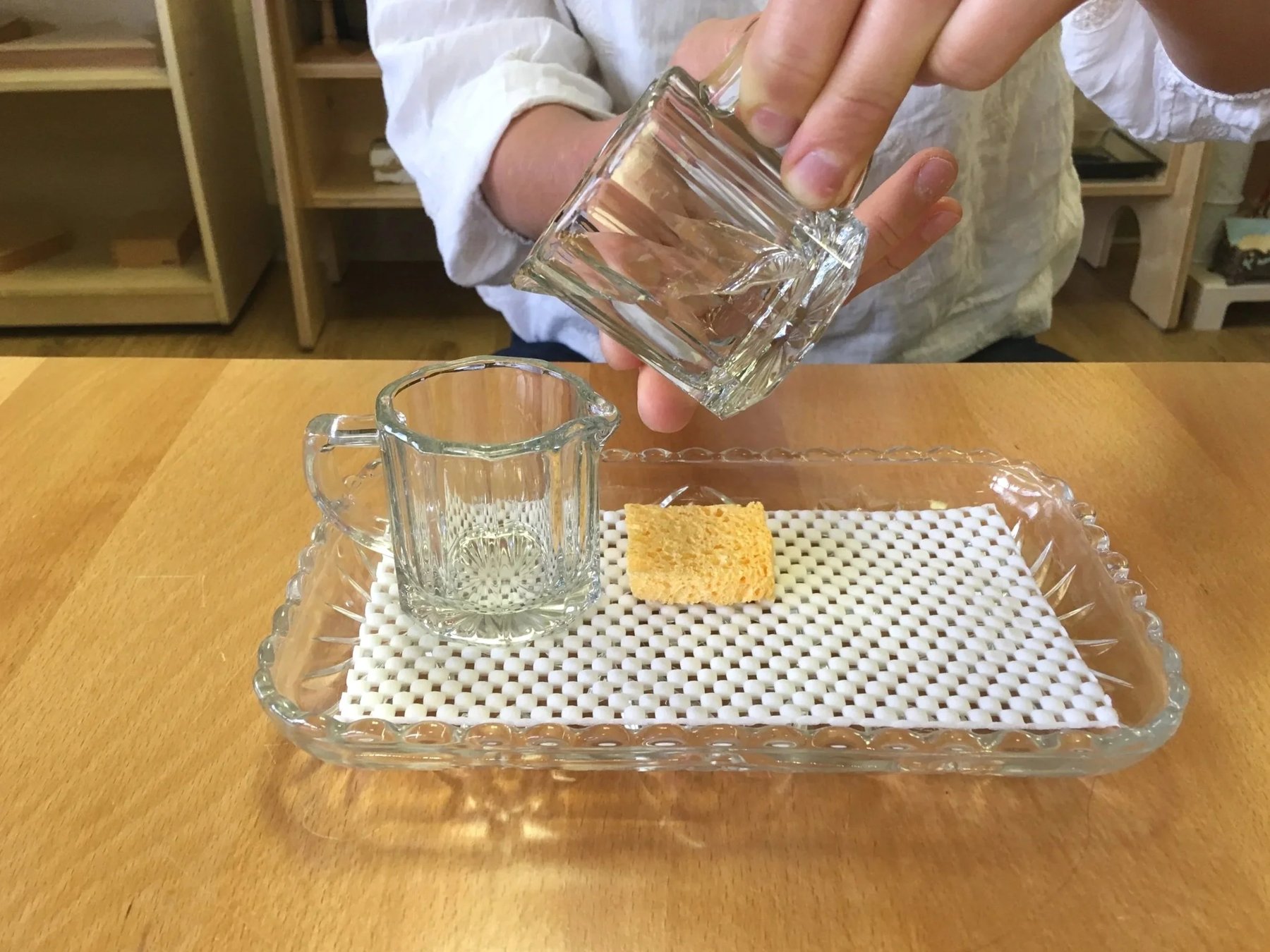 Close-up of a child's hands pouring water from one small glass pitcher into another over a tray during a Montessori pouring exercise